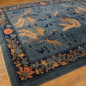 A dusty blue rug featuring tan and indigo phoenixes, with a detailed floral border, resting on warm wood floors.