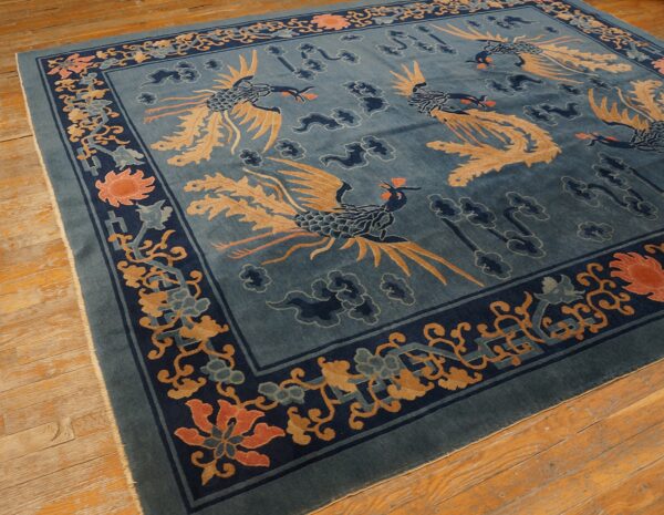 A dusty blue rug featuring tan and indigo phoenixes, with a detailed floral border, resting on warm wood floors.