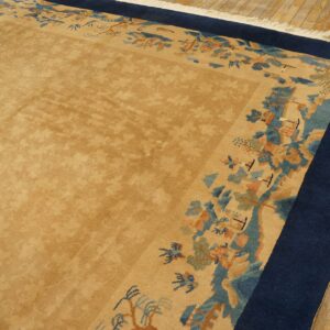 Muted tan rug with subtle overall pattern, framed by a blue border and multicolored scenic edge, on wood floors.