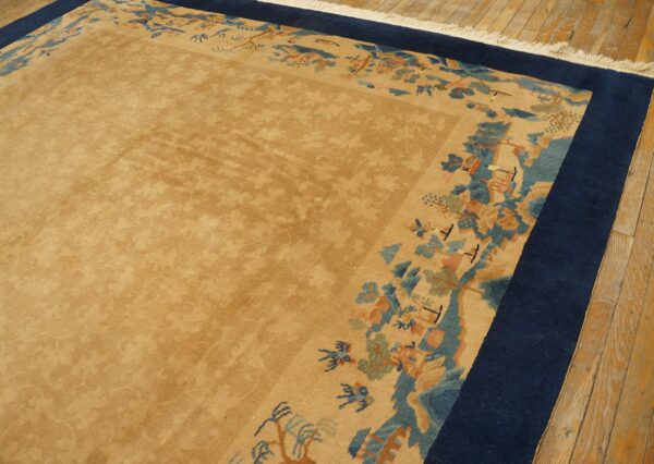 Muted tan rug with subtle overall pattern, framed by a blue border and multicolored scenic edge, on wood floors.