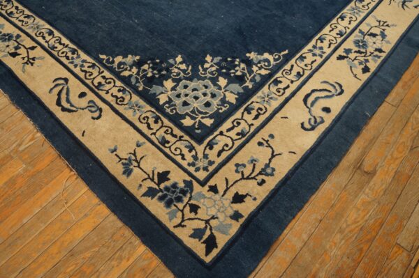The corner of a deep blue floral rug features a wide tan border on orange hardwood flooring.