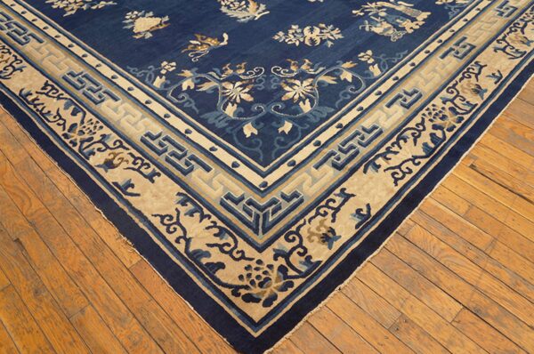 Corner view of a navy blue rug with scattered floral patterns and a wide beige geometric border on wood floors.