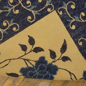 The edge of a dark blue scrolled rug is rolled, revealing a contrasting beige floral flatweave surface on wood.