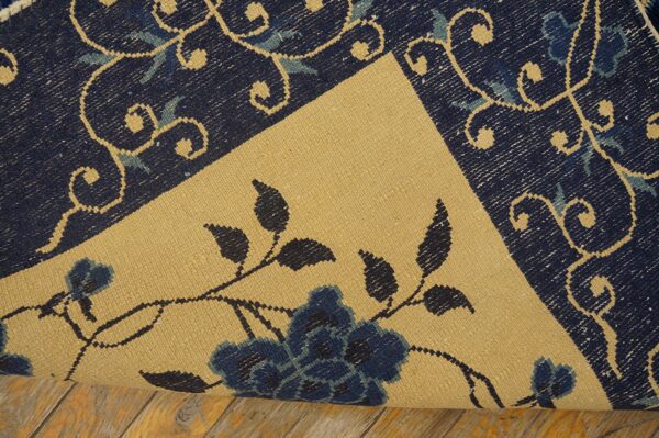 The edge of a dark blue scrolled rug is rolled, revealing a contrasting beige floral flatweave surface on wood.