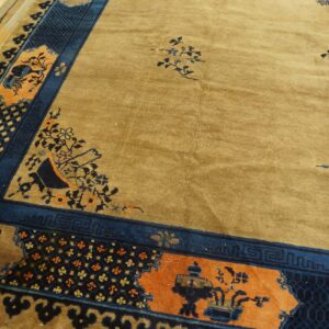 Gold-toned rug with dark blue, orange, and tan complex floral and geometric borders on plank flooring.