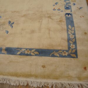 Tan high-pile rug with blue border and blue floral and vase motifs rests on distressed wood flooring.