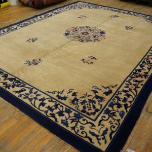 Large area rug, tan field with detailed navy blue floral borders, rests on worn wood planks.