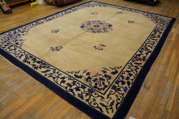 Large area rug, tan field with detailed navy blue floral borders, rests on worn wood planks.