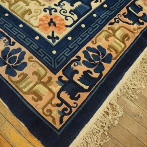Beige, navy, and orange patterned rug corner featuring fringes resting on distressed wood flooring.
