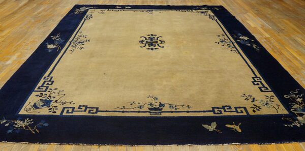 Tan rug with a dark blue border, floral motifs, and a central symbol displayed on wood flooring.