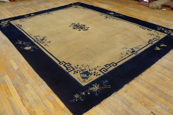 Large tan rug with a wide navy blue border featuring geometric key and delicate floral patterns on wood planks.
