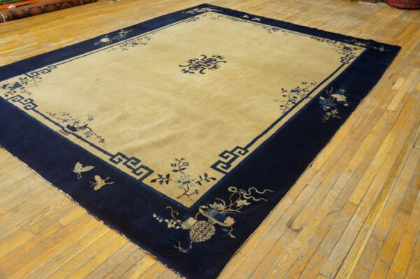Rectangular tan rug with a wide navy blue border featuring floral and key patterns on a wood floor.