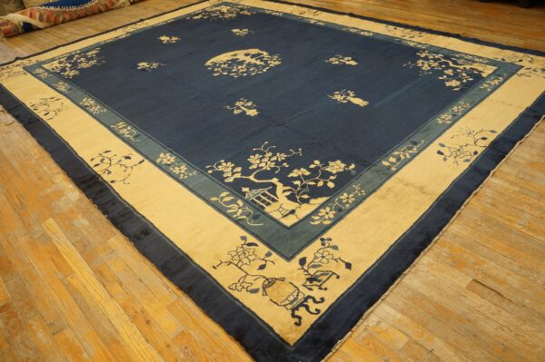 A large square rug with a navy blue field and beige/teal borders, featuring floral motifs on a worn wood floor.