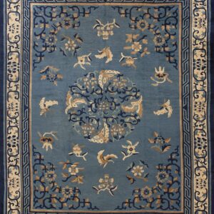 Dusty blue rug with stylized tan and navy floral patterns, framed by complex fretwork and vine borders.