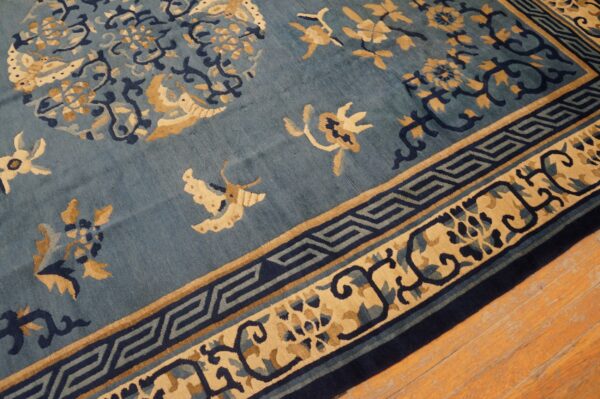 A blue rug featuring tan and navy floral motifs and key borders lies on light wood flooring.