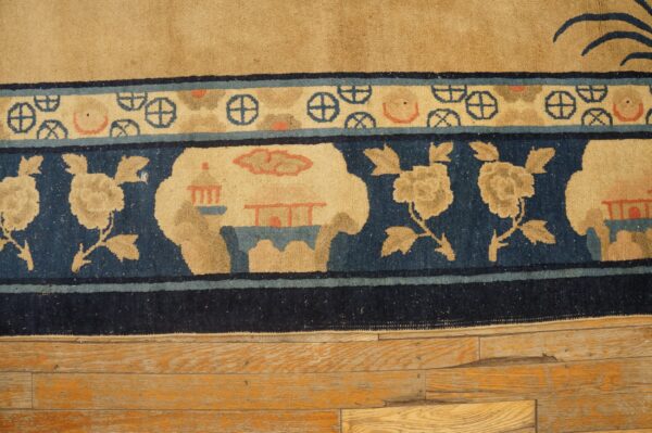 Tan rug with dark blue borders featuring flowers, structures, and wheel motifs on distressed wood plank flooring.