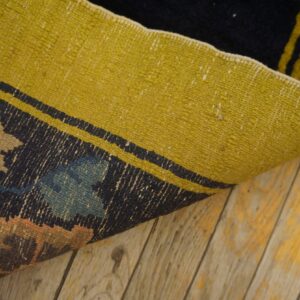 A curled rug displays a dark, patterned face with colorful motifs and a bright yellow reverse side on wood floors.