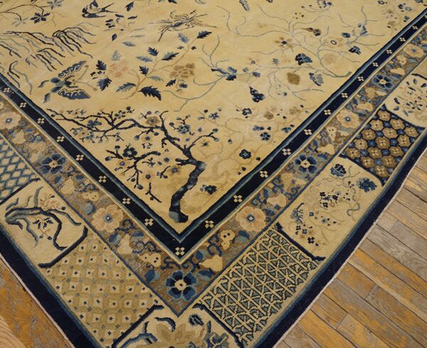 Pale yellow rug corner with a dense border and blue botanical motifs rests on distressed wood flooring.