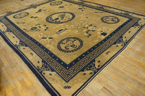 Golden tan rug with a navy blue greek key border and traditional medallions on wooden floorboards.