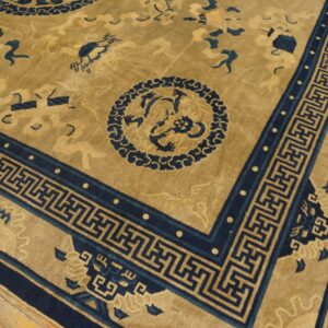 Close view of a low-pile rug with a tan field, dark blue motifs, and a geometric border on wood flooring.