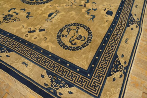 Close view of a low-pile rug with a tan field, dark blue motifs, and a geometric border on wood flooring.