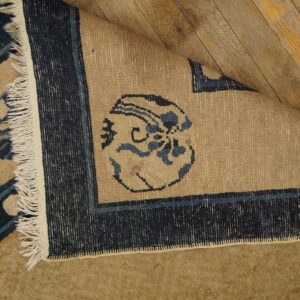 Beige and blue patterned rug corner folded back, revealing woven reverse side on yellow wood planks.