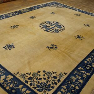 Square gold and blue rug featuring a central bird medallion and wide floral borders on distressed wood floors.