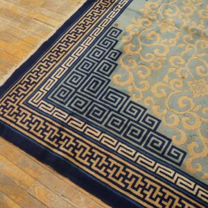 Teal rug with carved tan scrolling motifs and navy geometric borders shown on rustic wood flooring.