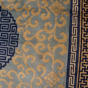 Close-up of a rug with blue, gold, and navy patterns, featuring scrolls and geometric borders on a wood floor.