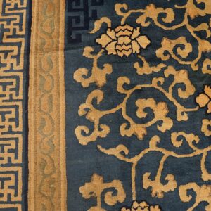Tan floral and scroll patterns cover the deep blue rug, bordered by a tan geometric meander design.