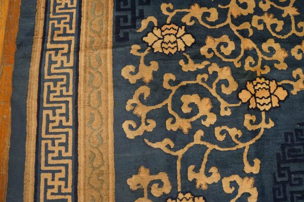 Tan floral and scroll patterns cover the deep blue rug, bordered by a tan geometric meander design.