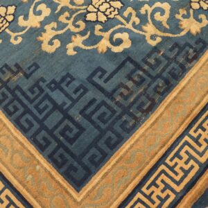 Low-pile teal and tan rug corner featuring floral designs and detailed geometric key patterns on wood flooring.