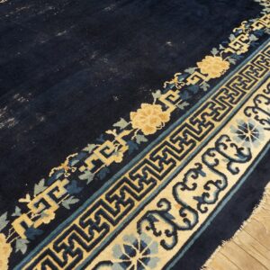 Dark blue rug corner shows gold and blue floral and geometric borders resting on light wood plank flooring.