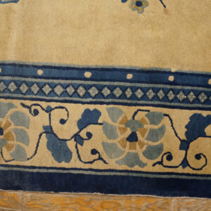 Tan rug close-up showing blue floral and geometric borders over rustic honey-toned wood floor.