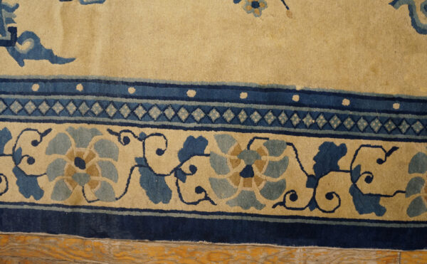 Tan rug close-up showing blue floral and geometric borders over rustic honey-toned wood floor.