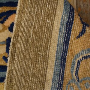 A folded rug corner reveals the woven khaki backing and the gold, blue, and beige pile on wood floors.