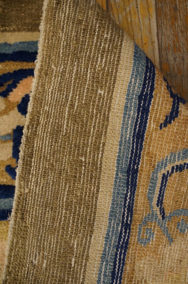A folded rug corner reveals the woven khaki backing and the gold, blue, and beige pile on wood floors.