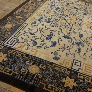 Low-pile rug with a cream field, blue floral vines, and a dark geometric border on distressed wood floors.