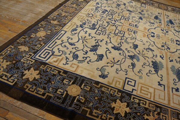 Low-pile rug with a cream field, blue floral vines, and a dark geometric border on distressed wood floors.