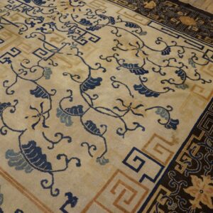 Cream and indigo floral rug with a complex dark border rests on rustic wooden flooring.