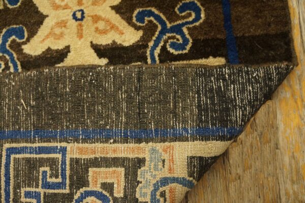 Dark brown, tan, and blue patterned rug corner folded to display the low pile backing on wood flooring.