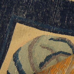 Close-up of a rug corner turned up, showing a dark blue, tan, and sage green pattern over wood flooring.