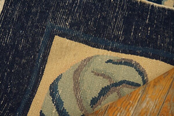 Close-up of a rug corner turned up, showing a dark blue, tan, and sage green pattern over wood flooring.