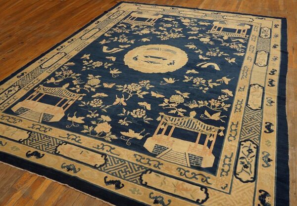 A large navy blue rug with a light beige scenic pattern featuring pavilions, flowers, and a medallion, lying on a wooden floor.
