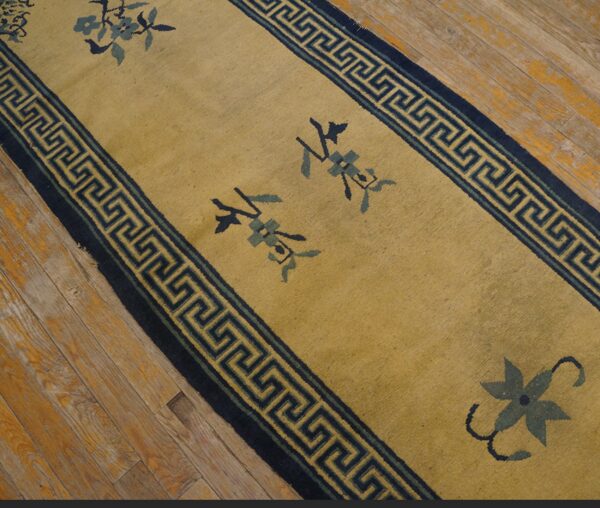 Gold runner rug with navy greek key borders and stylized blue floral motifs lies on distressed wood planks.