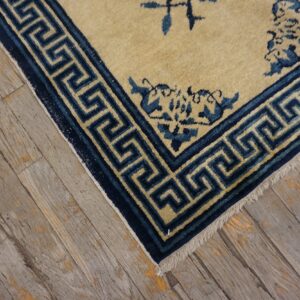 Tan rug with dark blue floral and Greek Key motifs rests on weathered gray hardwood planks.