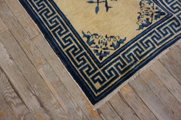 Tan rug with dark blue floral and greek key motifs rests on weathered gray hardwood planks.