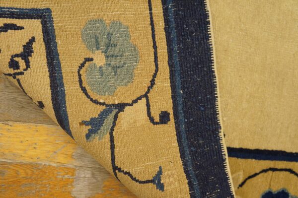 Tan, low-pile rug with navy and light blue floral designs rolled up on distressed wood flooring.