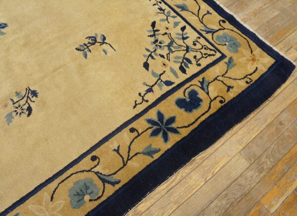 Close-up of a camel rug with scattered blue floral sprays and a navy blue border on rustic wood planks.