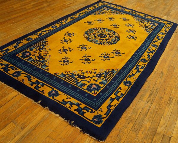 Gold rug featuring blue floral patterns and geometric borders rests on diagonal wooden plank flooring.